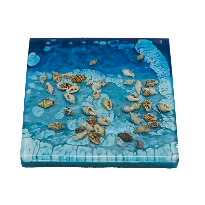 A closeup of a single ocean-inspired square coaster with shells embedded in the resin in which is the material it is made from.