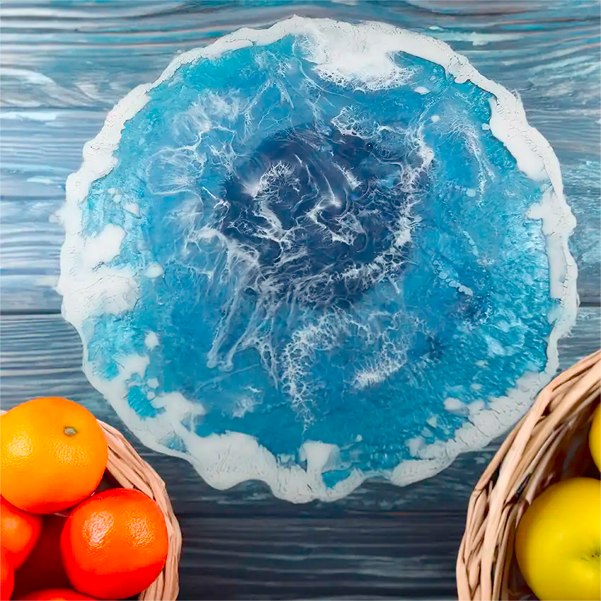 The ocean inspired trivet or centerpiece, resting on a table next to fruit baskets, has geode shaped edges, measures 13 inches round, and is made of epoxy resin.