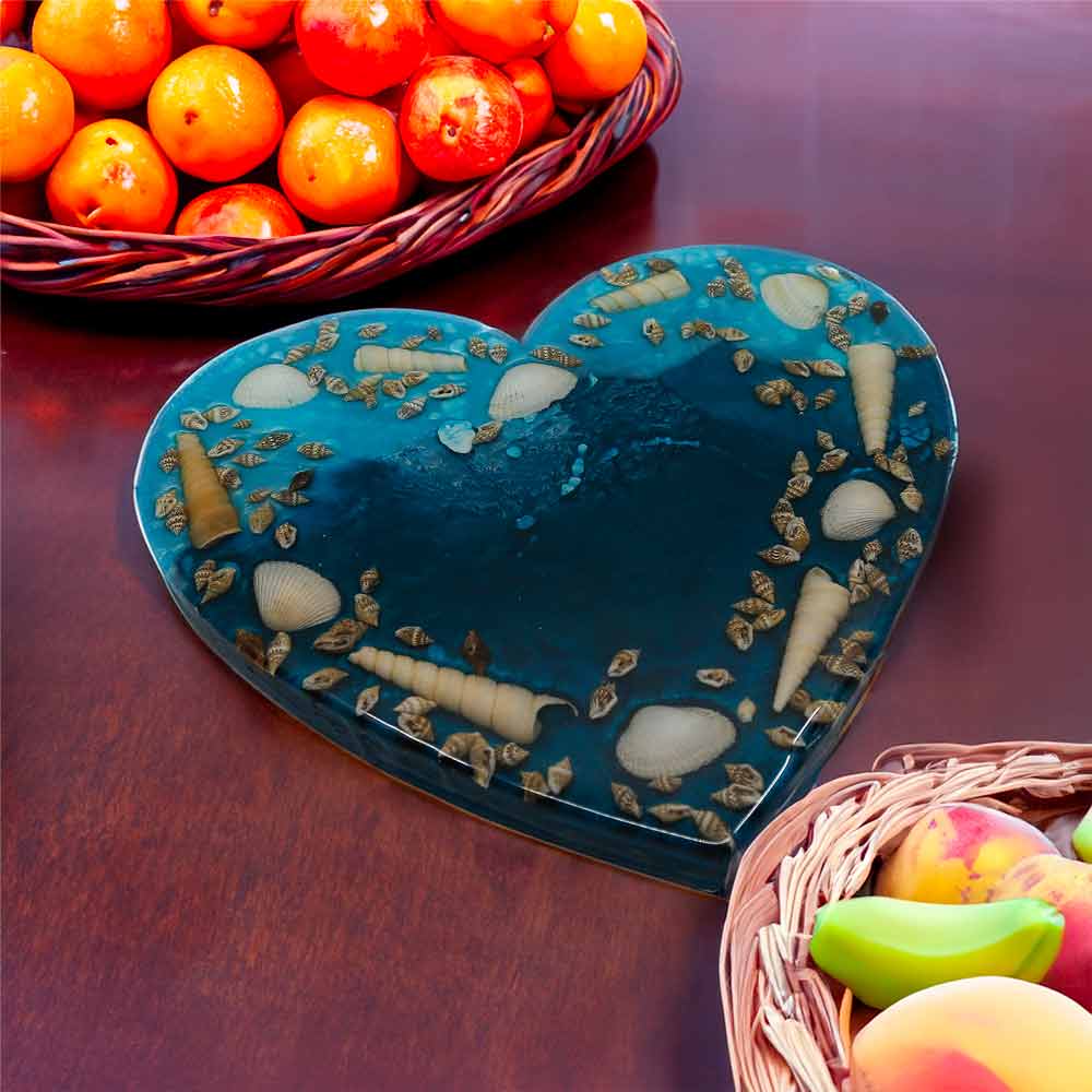 The resin, ocean-inspired, heart-shaped trivet with shells embedded sits on a brown table next to a basket of fruit.