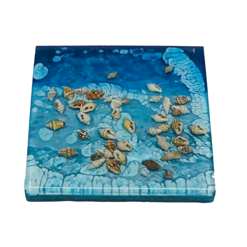 A closeup of a single ocean-inspired square coaster with shells embedded in the resin in which is the material it is made from. 