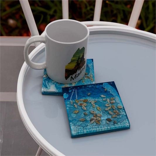 A pair of square ocean themed coasters with real shells rests on a small table, one with a coffee cup on it. 